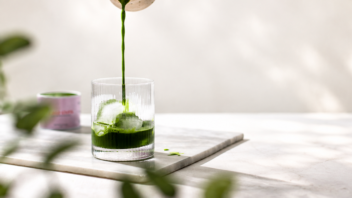 A clear glass tumbler with a green patterned base sits on a light stone tray, with a dark green liquid being poured into it from above. The scene is softly lit with blurred green leaves in the foreground and a bright neutral background.
