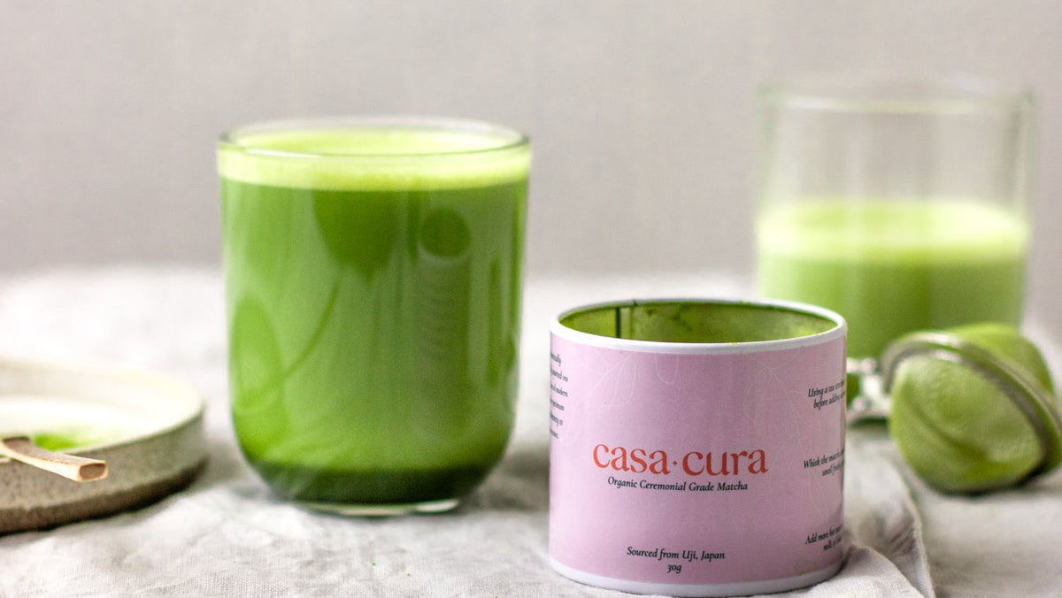 A pink canister of Casa Cura Organic Matcha sits on a textured light gray cloth next to two glasses filled with bright green tea, with a lime slice in the background.